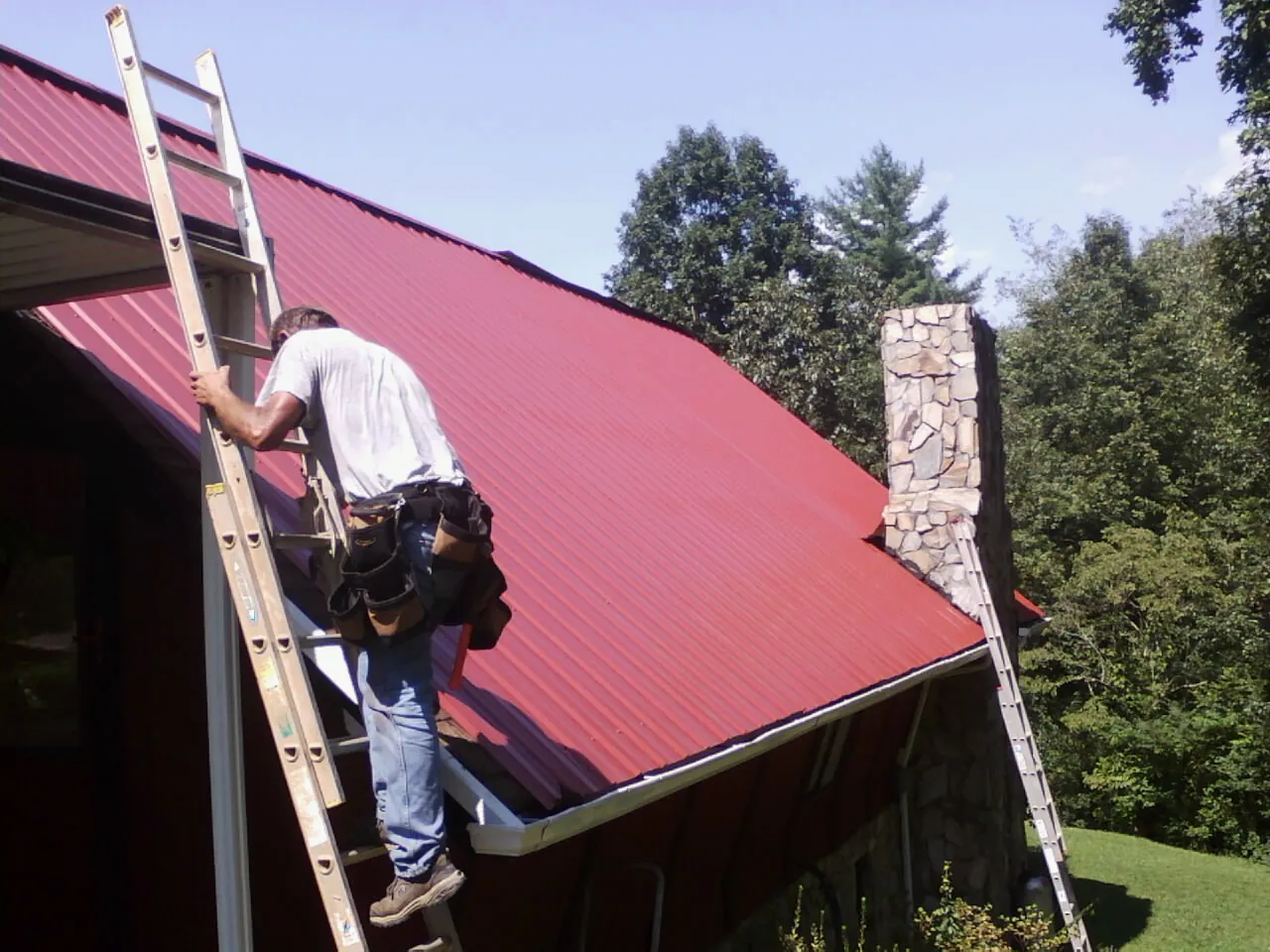 Roofer on a ladder beside a red metal roof and stone chimney — Soffit & Fascia Repair by Roofing Brothers in Hennepin